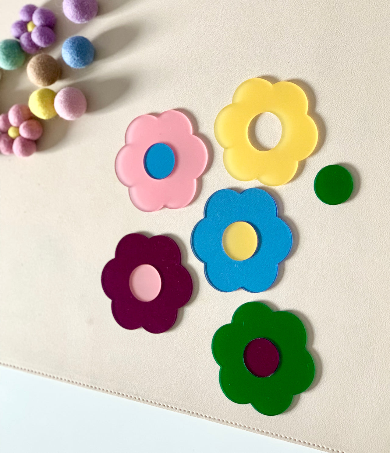 Five colorful flower-shaped acrylic cutouts in pink, yellow, blue, purple, and green on a light surface near felt balls
