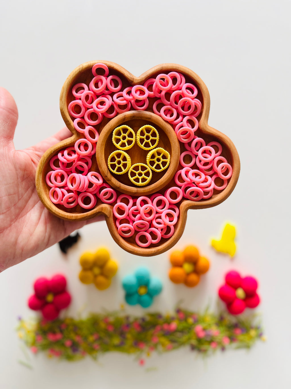 Hand holding wooden flower-shaped tray filled with pink rings and yellow wheel-shaped pasta on white background