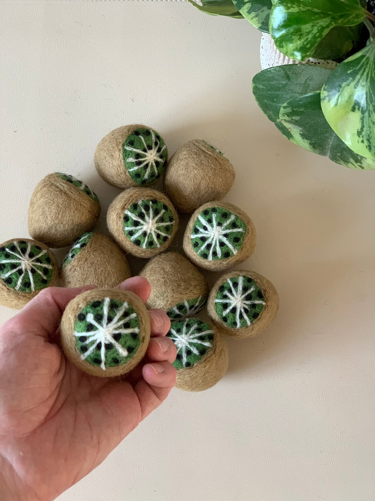 Felted wool balls designed to resemble kiwi fruit halves arranged on a light surface near green houseplant leaves