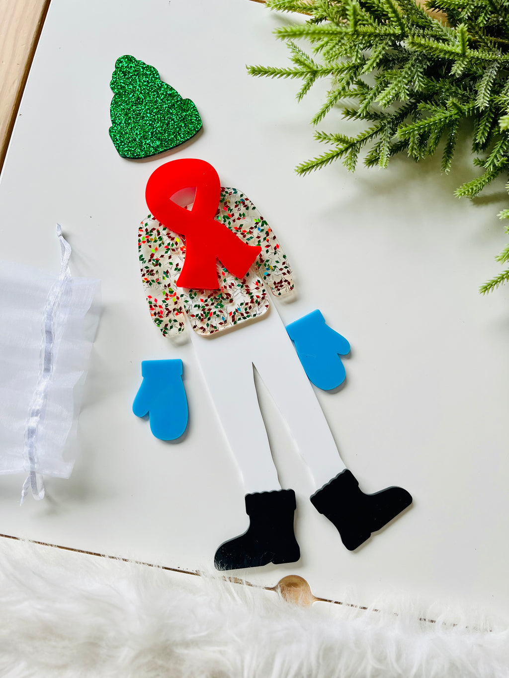 Cutout acrylic figure wearing glittery green hat, floral top with red scarf, blue mittens, white pants, and black boots on white surface