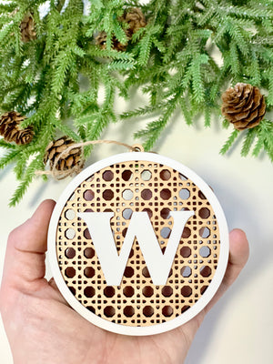 Hand holding round rattan Christmas ornament with white border and letter W in center, pine branches and pinecones background