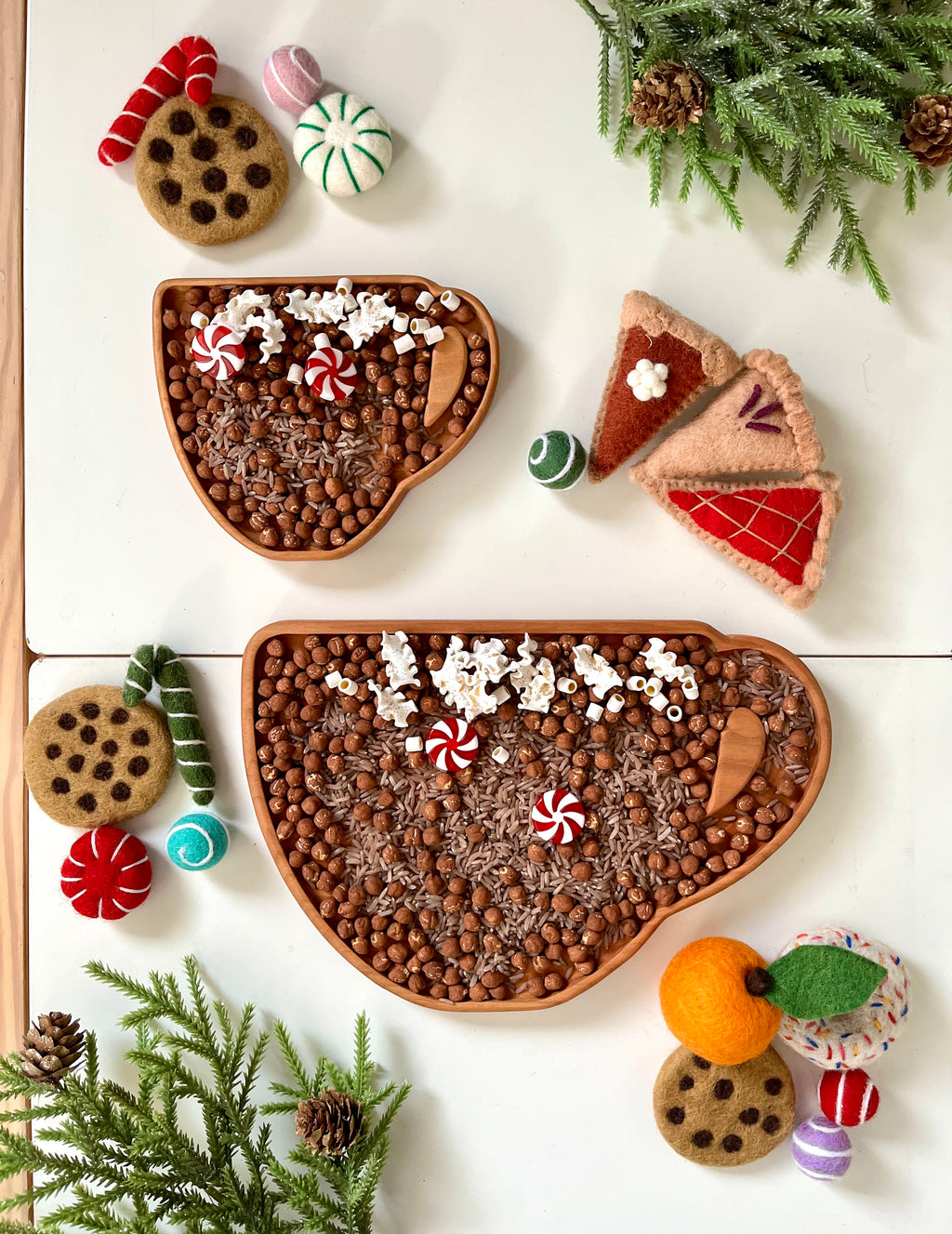 Two wooden cup-shaped trays filled with brown beads, felt peppermint candies, popcorn, and wooden scoops on white surface with felt cookies and pine branches