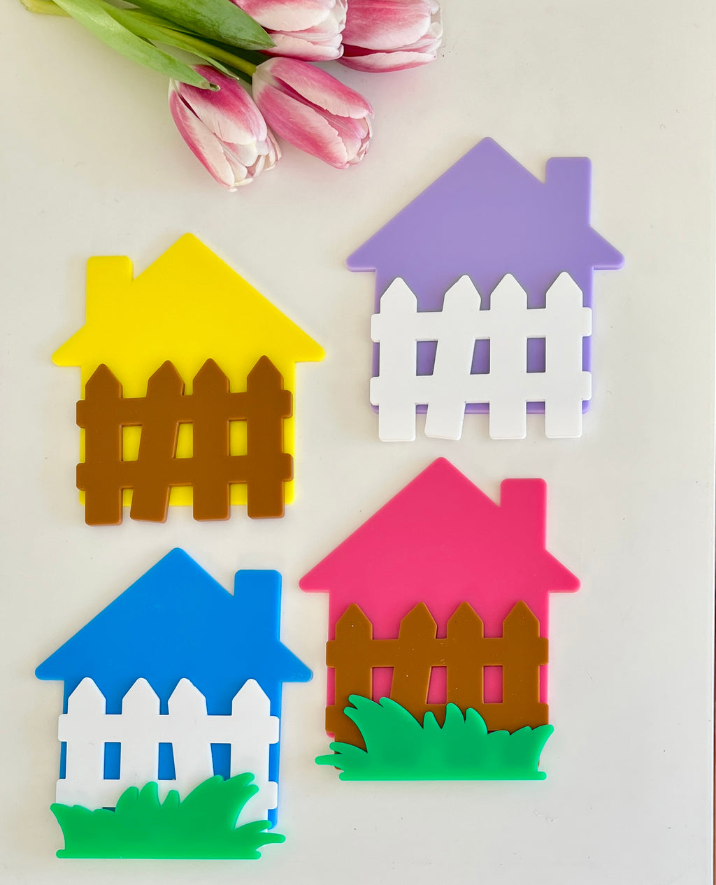 Four colorful house-shaped cutouts with white or brown fences and green grass pieces beside pink tulips on beige surface