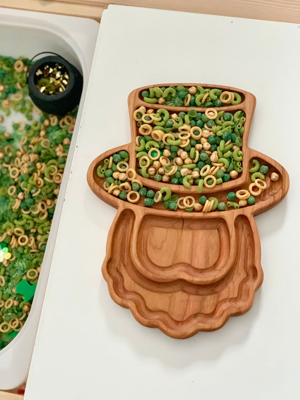 Wooden tray shaped like leprechaun hat and beard filled with green and beige cereal rings and beads on white surface
