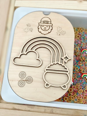 Wooden board with engraved leprechaun, shamrocks, rainbow, cloud, pot of gold, and coin designs above colorful ring shapes