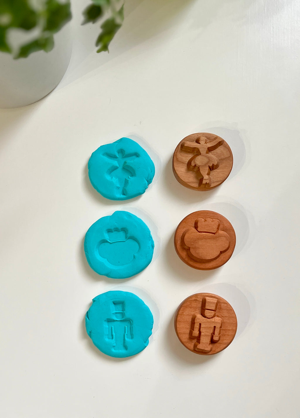 Three wooden stamps with engraved figures and their blue clay imprints on a white surface near a green plant pot