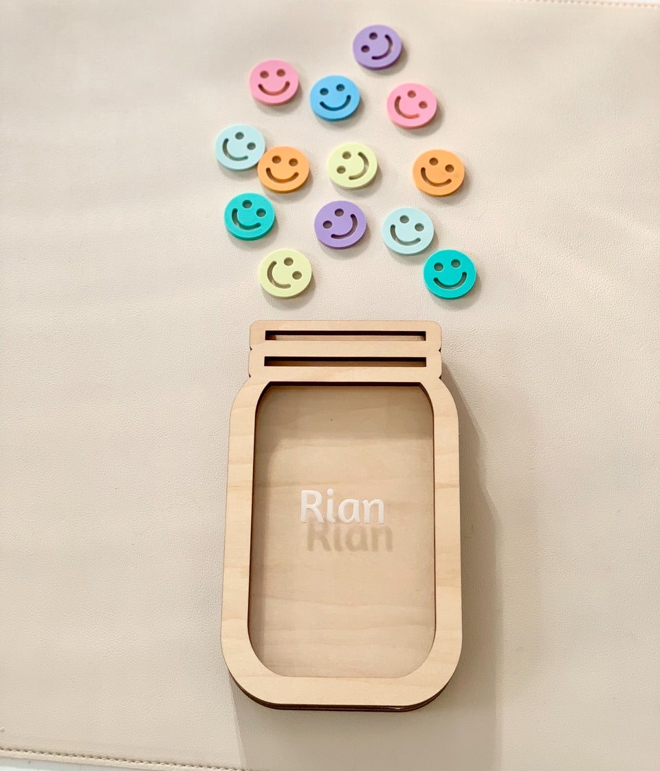 Wooden personalized reward jar with engraved name and colorful smiley face tokens on light background