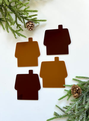 Four brown house-shaped acrylic coasters in varying shades with pinecones and evergreen branches nearby