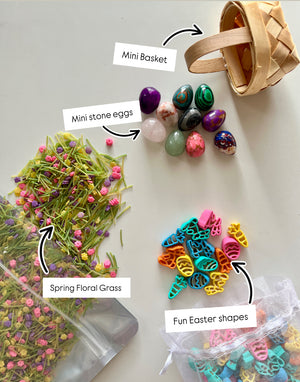Small woven basket, smooth colorful stone eggs, pastel plastic Easter shapes, and green floral grass on white background