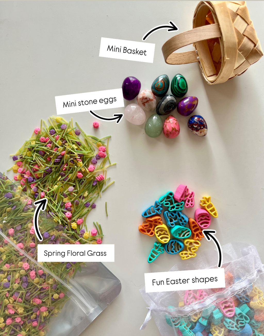 Small woven basket, smooth colorful stone eggs, pastel plastic Easter shapes, and green floral grass on white background