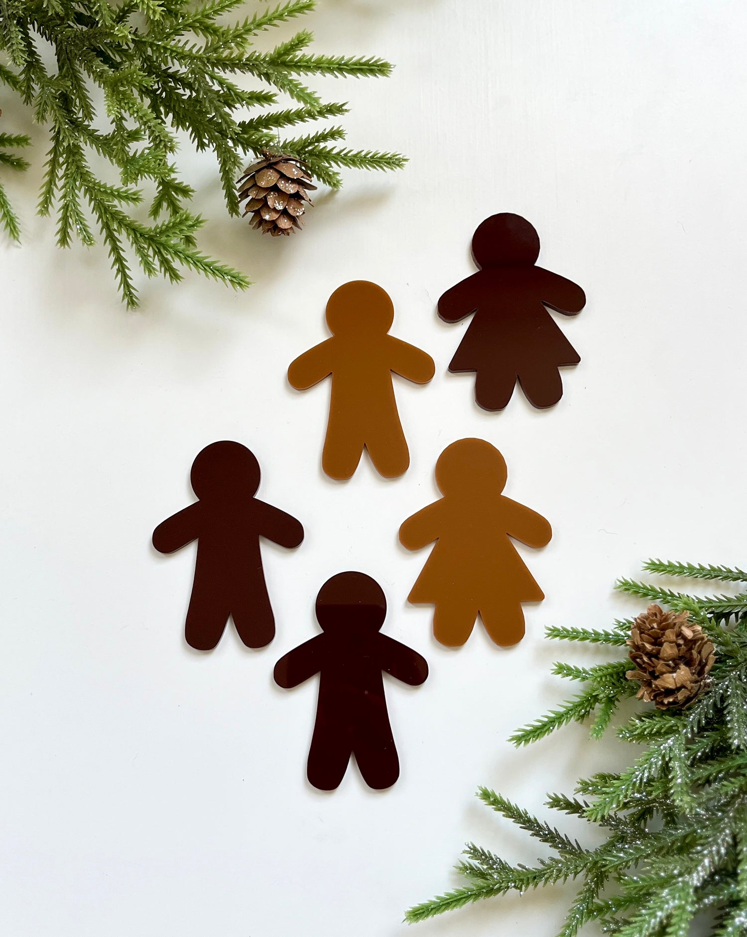 Five gingerbread-shaped dry erase people in varying brown tones surrounded by pine branches and pinecones on white background
