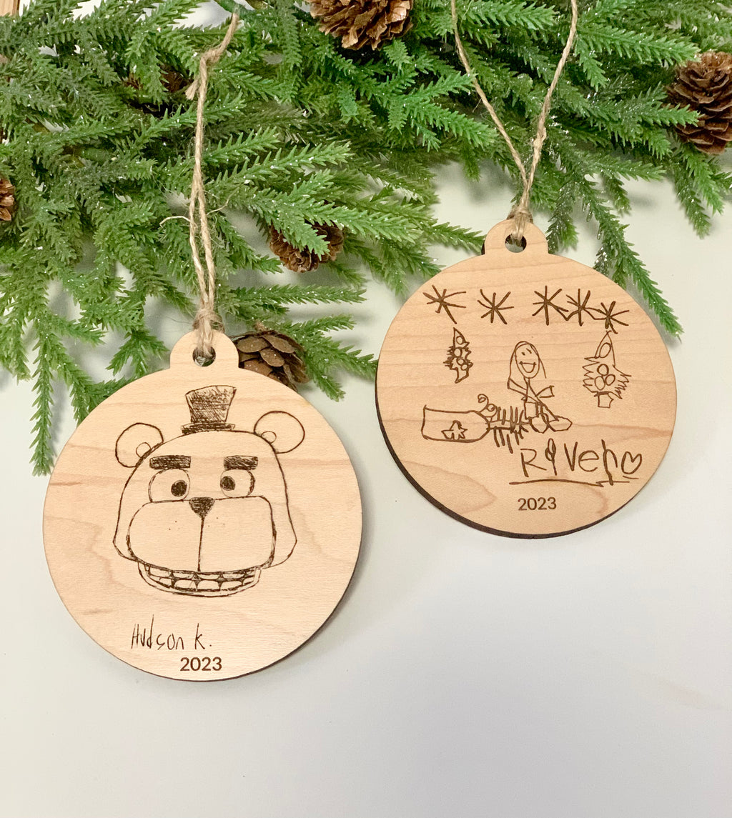 Two round wooden Christmas ornaments engraved with childlike drawings and names hanging from twine on a white surface with pine branches above