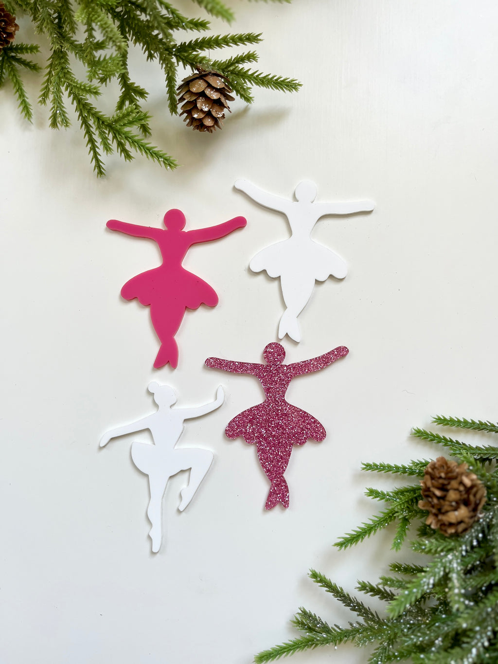 Four ballerina-shaped cutouts in pink, white, and glittery pink surrounded by pine branches and pinecones on white background
