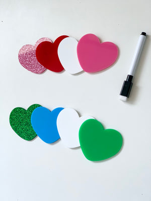 Sets of colorful heart-shaped dry erase boards in pink, red, white, blue, green, and glitter with a dry erase marker on white surface