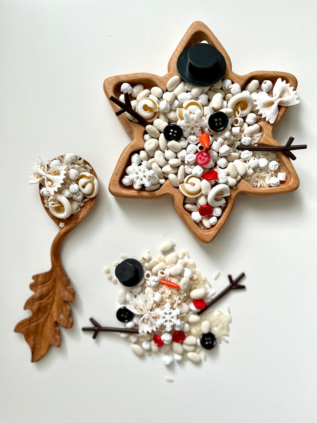 Wooden star-shaped tray filled with white beans, buttons, red beads, and black twig-like decorations next to a leaf-shaped spoon and scattered contents on white surface