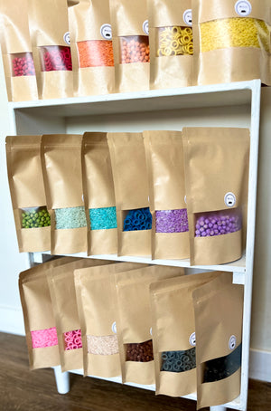 Multiple brown kraft paper bags filled with assorted brightly colored sensory filler types arranged on white wooden shelves