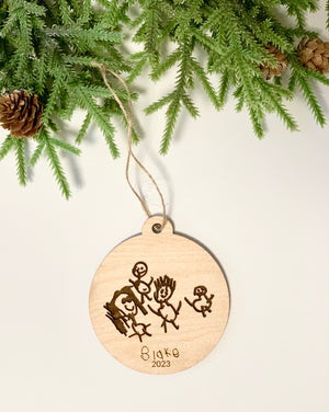 Round wooden Christmas ornament with childlike family drawing and "Blake 2023" engraved hanging from twine near pine branches and cones