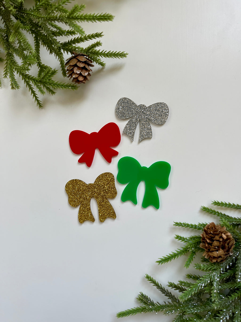 Four decorative bows in red, green, silver glitter, and gold glitter on white surface with pinecones and greenery