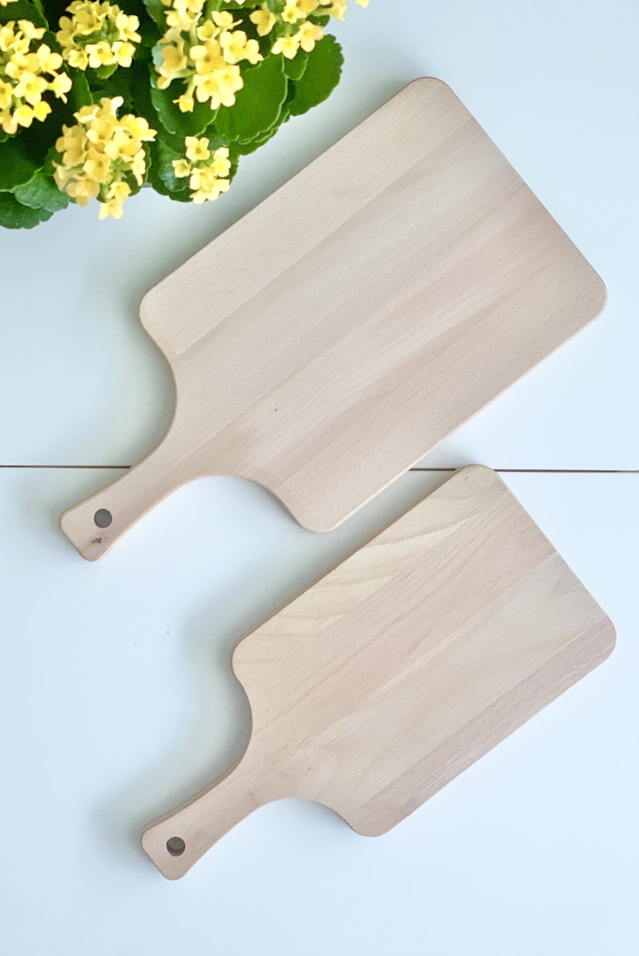 Two rectangular light wood cutting boards with handles placed on a white surface next to a yellow flowering plant