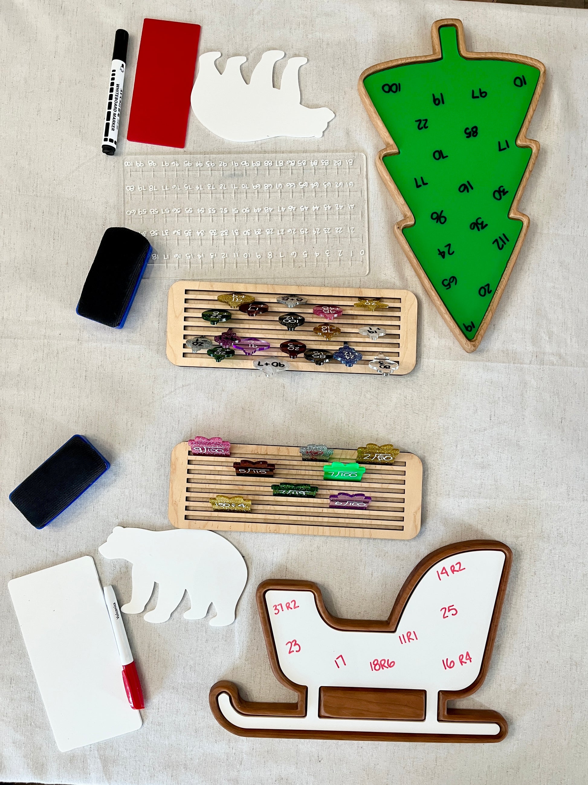 Wooden sled-shaped sensory tray with whiteboard markers, erasers, polar bear shapes, and green Christmas tree tray on white surface