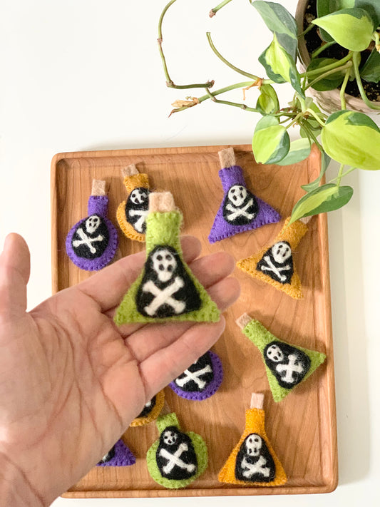 Hand holding green felt potion bottle with skull and crossbones on wooden tray with more bottles and plant nearby