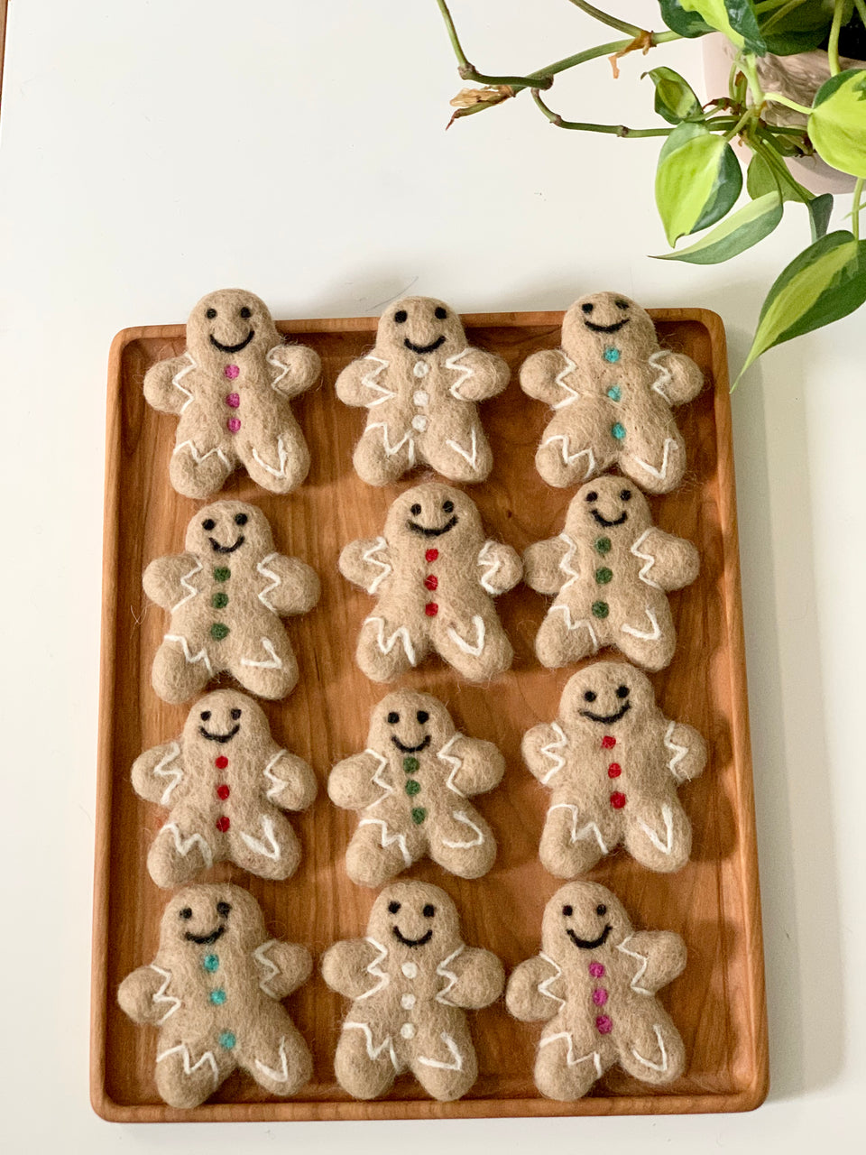 Twelve handmade felt gingerbread people with colorful button details on wooden tray beside green plant on white background
