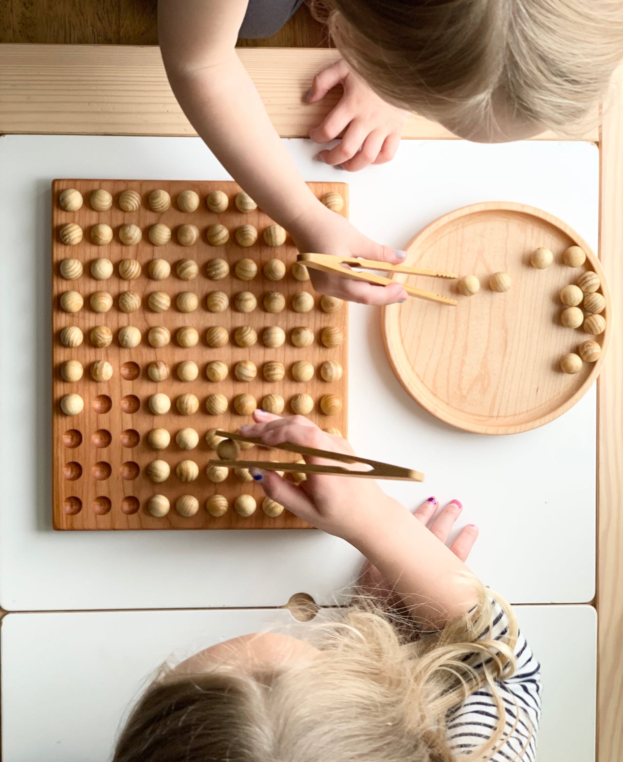 Children using wooden tweezers to place natural wood balls into square hundred sorting board and round tray on white table