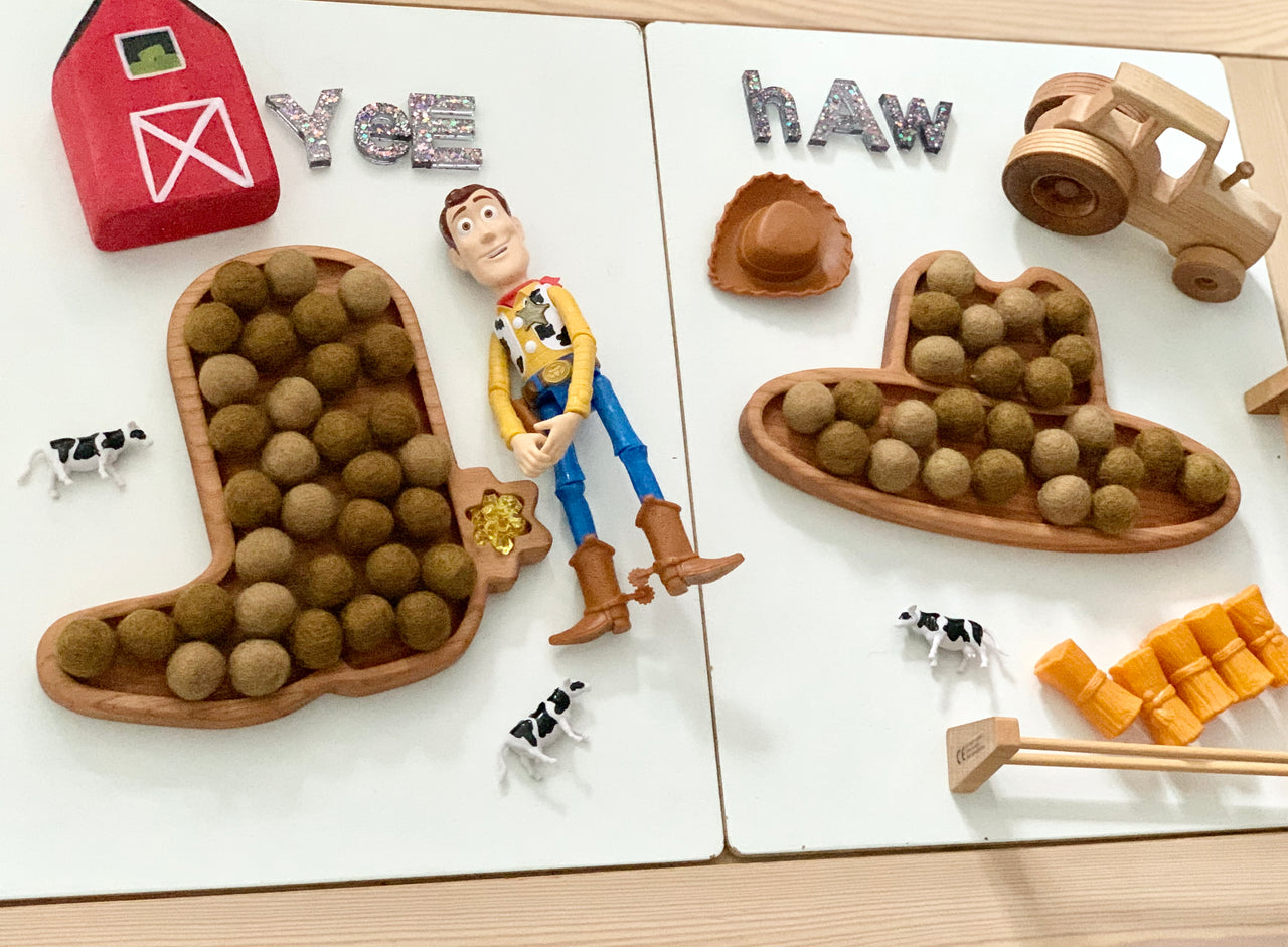 Wooden cowboy boot and hat sensory trays filled with felt balls, toy barn, Woody figure, cows, and wooden farm tractor on white table