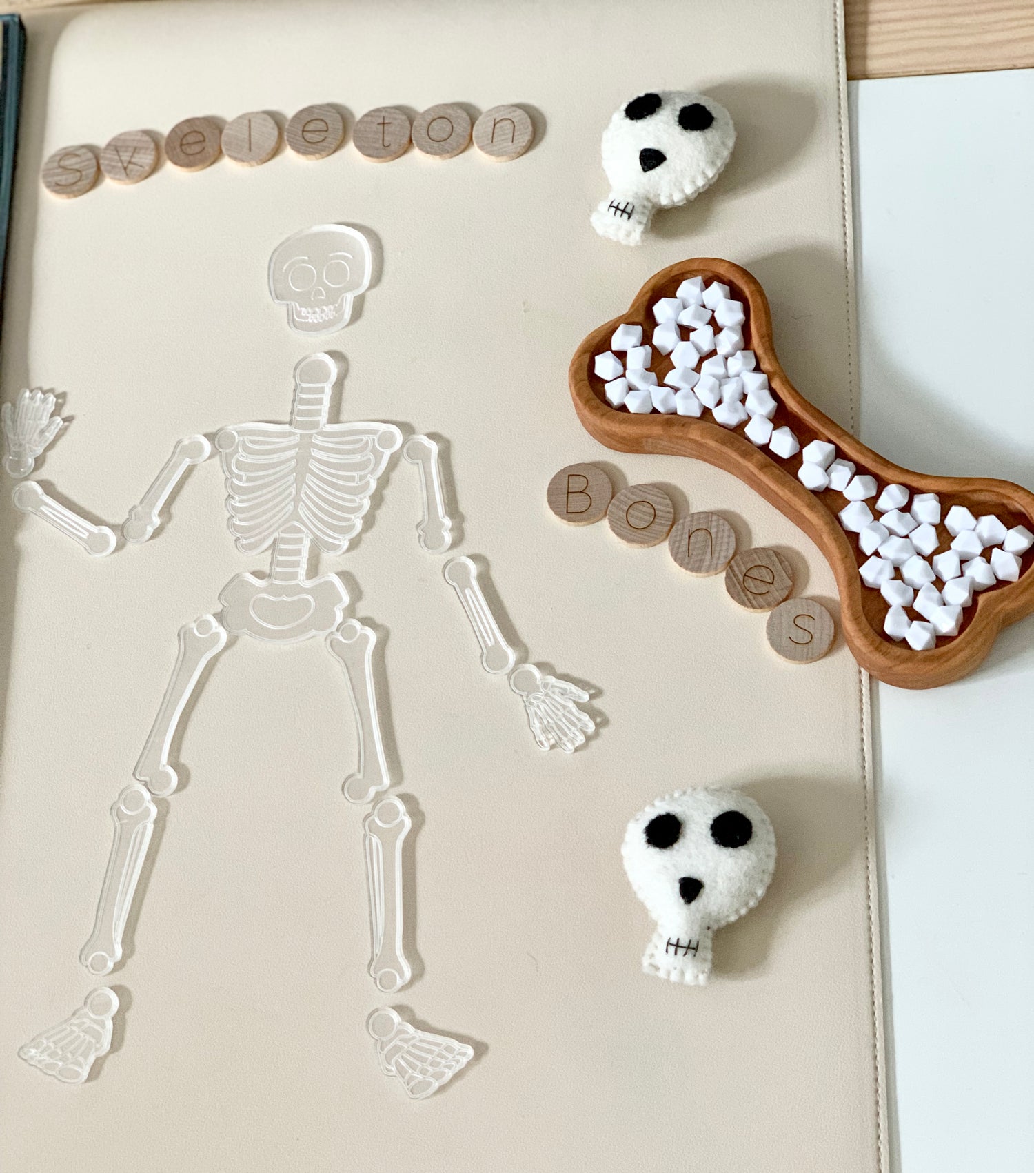 Clear skeleton puzzle with separate bone pieces on white mat, surrounded by wooden letter tiles and felt skulls