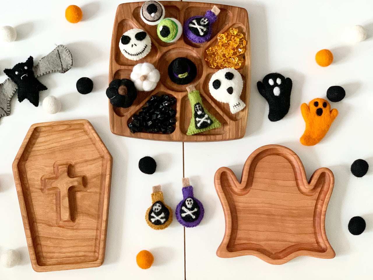 Wooden coffin-shaped and ghost-shaped sensory trays with felt Halloween-themed toys and scattered felt balls on white surface