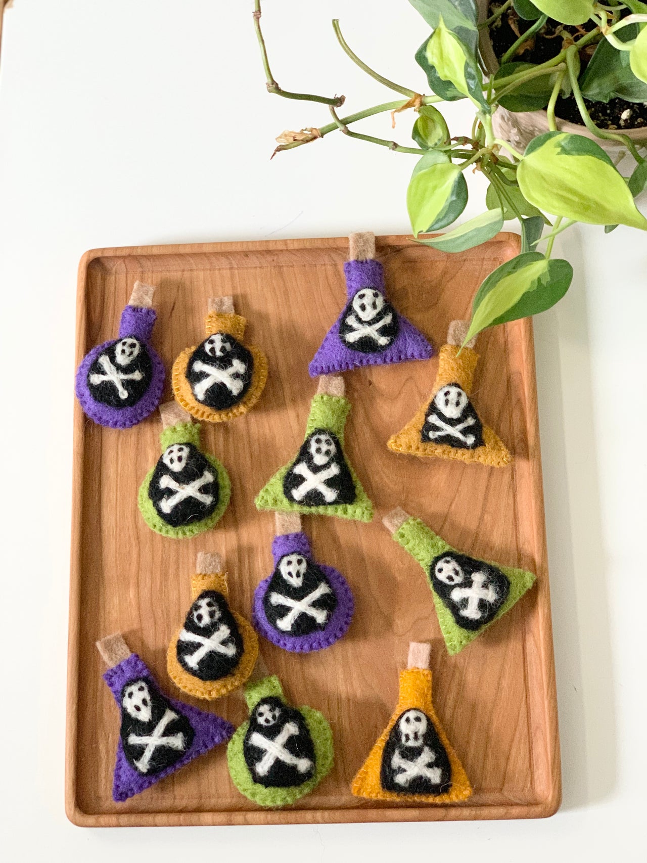 Handmade felt potion bottles in purple, orange, and green with skull and crossbones designs on wooden tray with plant nearby