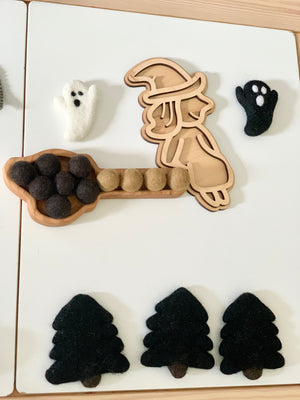 Wooden witch-shaped sensory tray with broom filled with felt balls, black and white ghost toys, and black felt trees on white surface