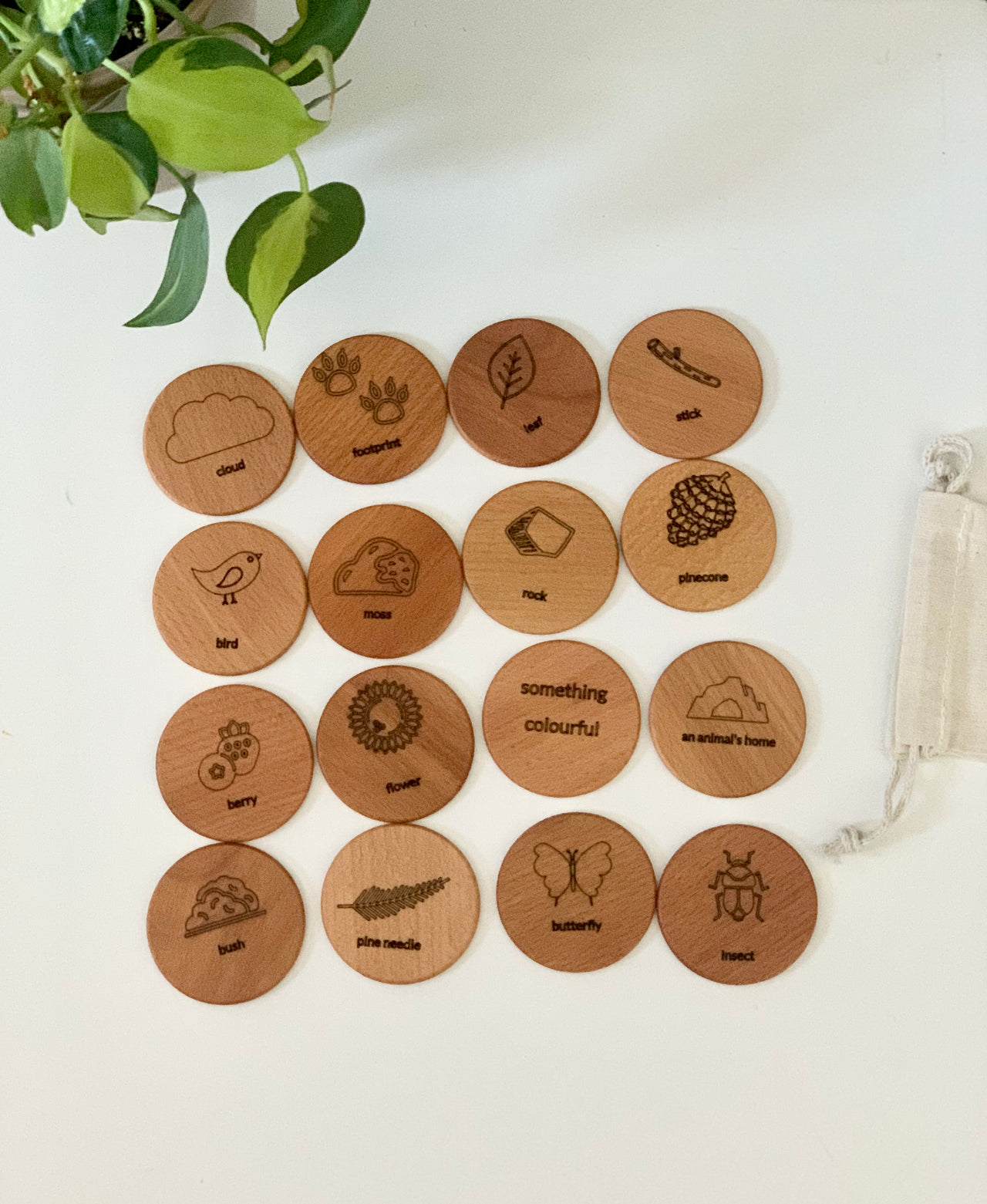 Wooden scavenger hunt coins engraved with nature-themed icons and words, displayed next to a drawstring bag on white surface