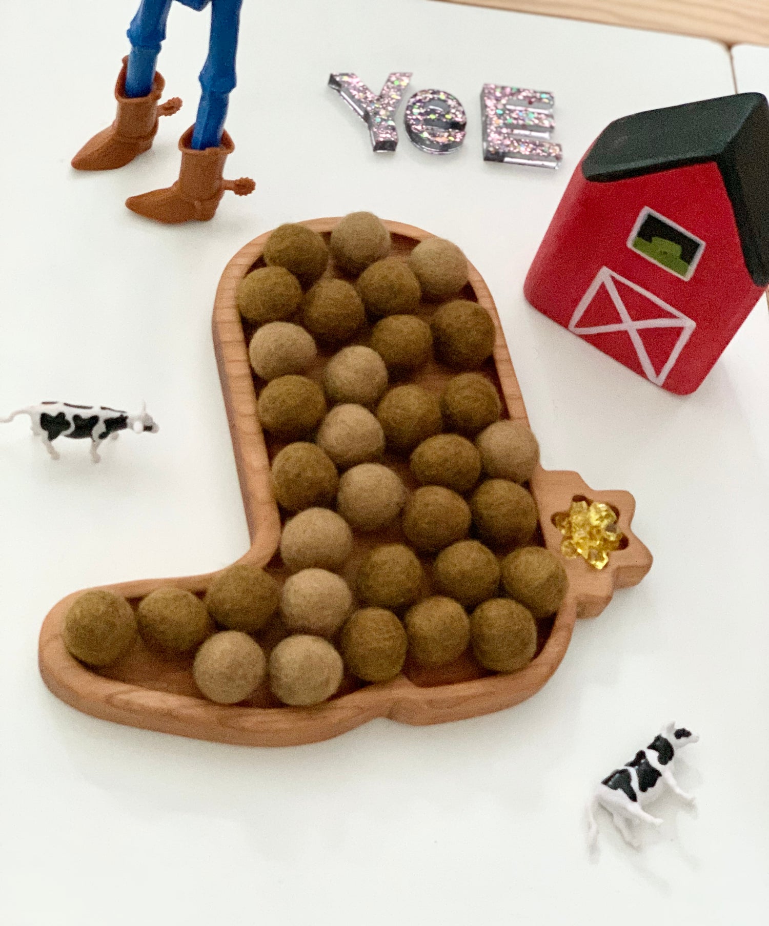 Wooden cowboy boot-shaped sensory tray filled with brown felt balls surrounded by toy barn, cows, and figure with boots on white table