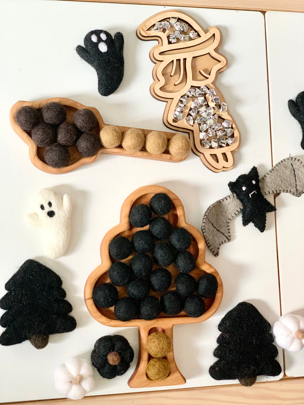 Wooden tree-shaped sensory trays filled with black and brown felt balls surrounded by felt ghost, bat, tree, and pumpkin decorations on white table