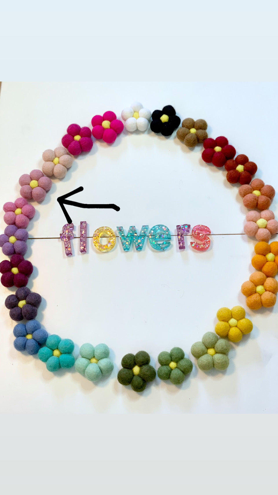 Single handmade felt flower in pale pink and yellow among multicolored felt flowers arranged in a circle on white background