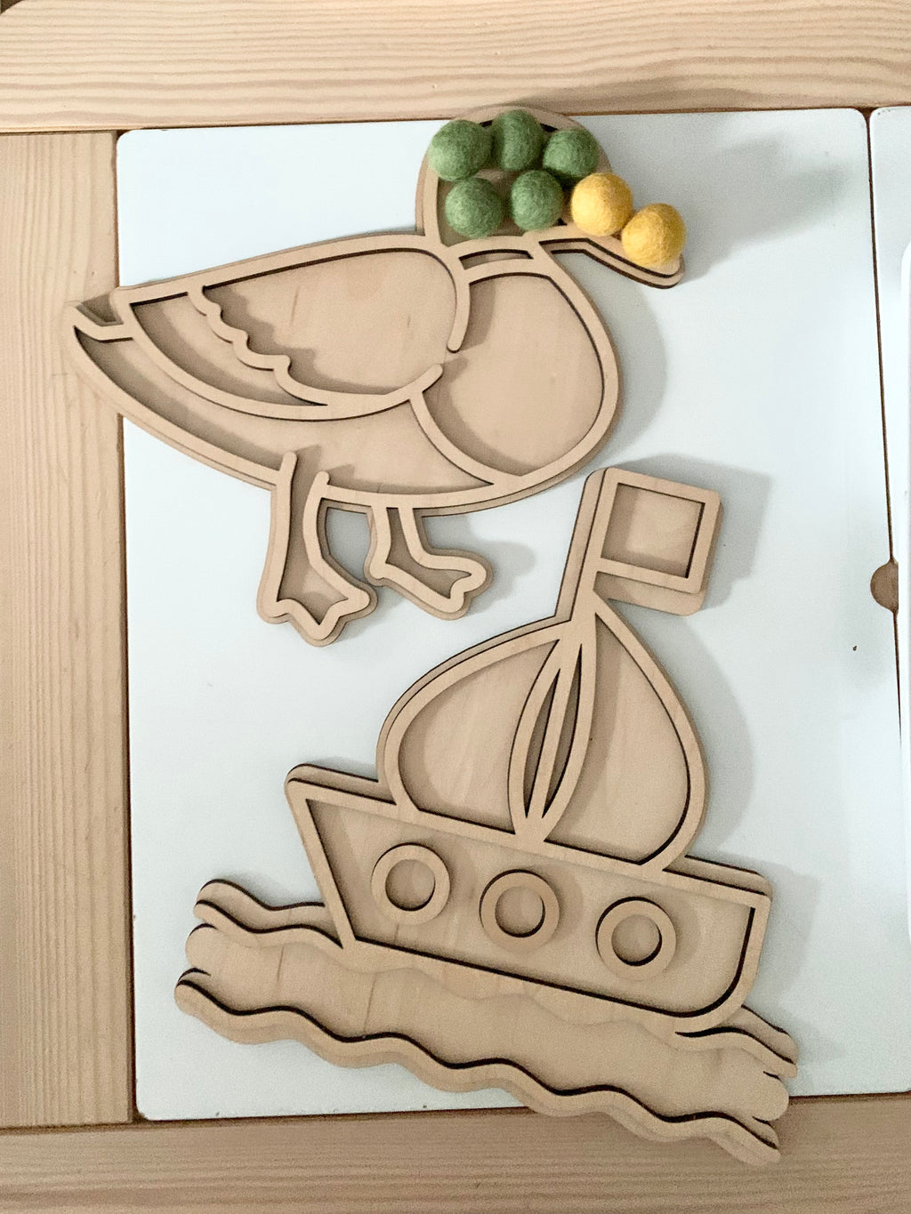 Fillable duck-shaped play tray with green and yellow felt balls and wooden sailboat tray on white surface