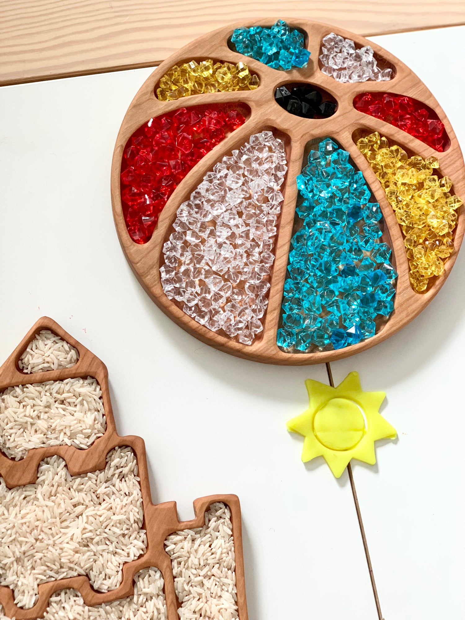 Wooden beach ball sensory tray filled with colorful translucent beads and a yellow sun-shaped toy on white surface