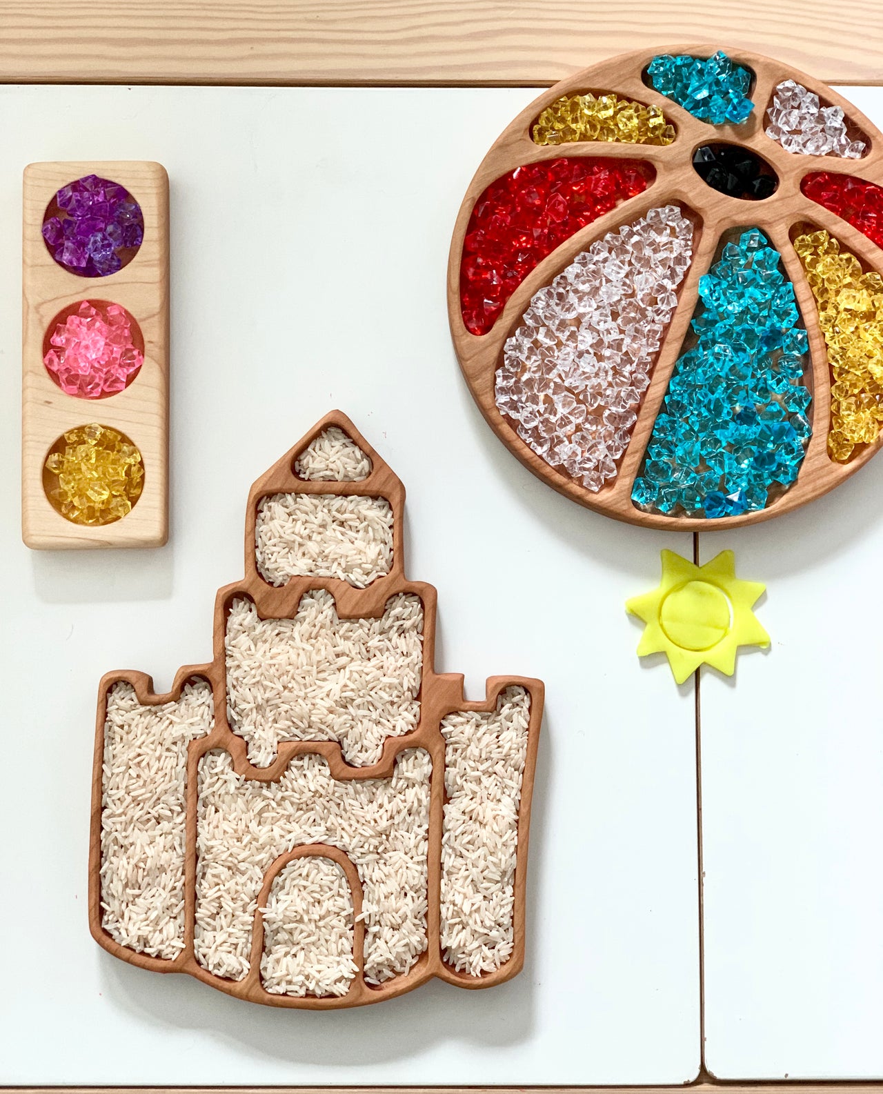 Wooden castle-shaped sensory tray filled with rice next to color sorting tray and wooden block with translucent gems on white table