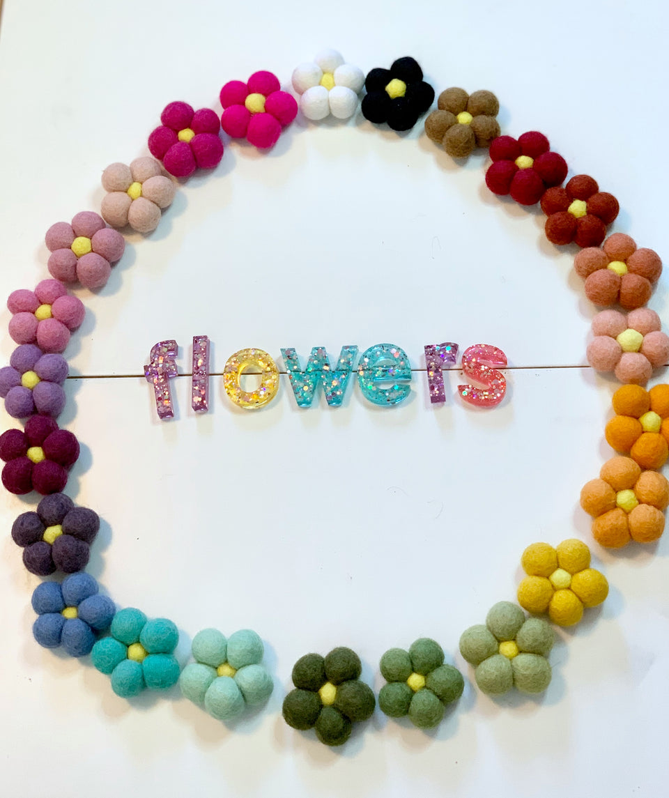 Circle of colorful felt wool flowers surrounding glittery resin letters spelling flowers on white background