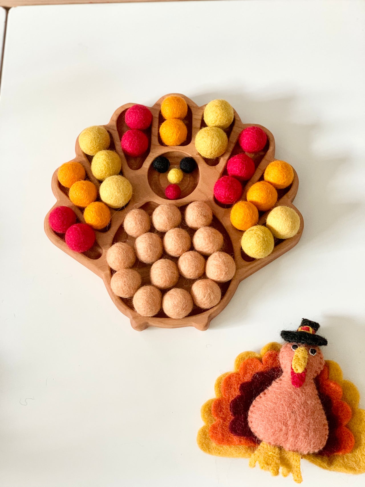 Wooden turkey-shaped sensory tray filled with orange, yellow, red, and brown felt balls on white table with felt turkey craft