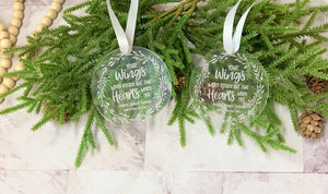 Two round clear acrylic memorial Christmas ornaments with white engraved text and decorative leaves hanging on green pine branches