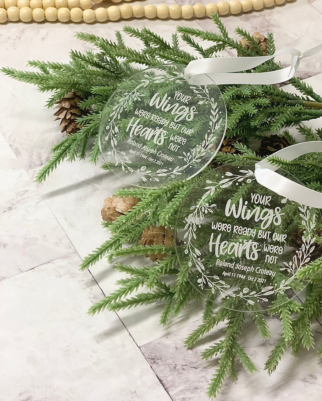 Clear round acrylic memorial Christmas ornaments engraved with white script and floral design, resting on pine branches
