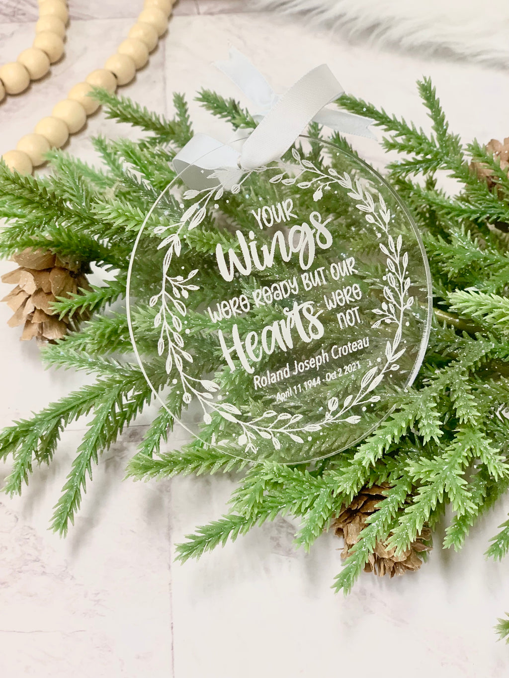 Clear round acrylic memorial Christmas ornament with white engraved wreath and personalized text on greenery background