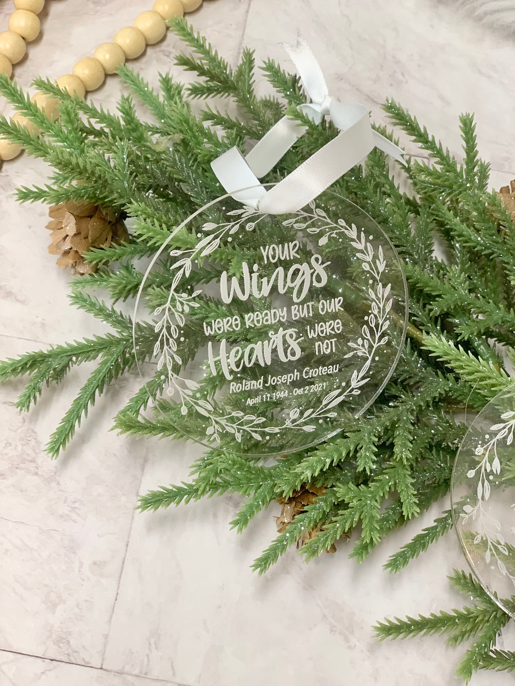 Round clear acrylic memorial Christmas ornament with white engraved script and leaf wreath design on greenery background