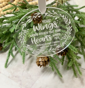 Round clear acrylic memorial Christmas ornament with engraved white floral wreath and sentimental text hanging on evergreen branch