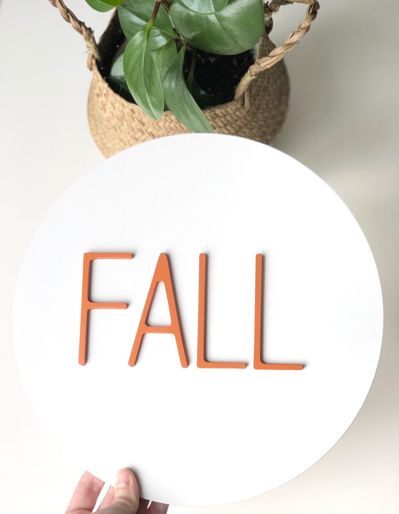 Round white sign with 3D orange "FALL" letters made from layered MDF held in front of a potted plant