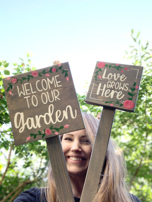 Small square wooden garden signs with white cursive text and pink floral accents held outdoors by a smiling woman