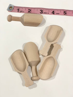 Five natural wooden sensory bin scoops and scoop with handle next to pink measuring tape on white surface