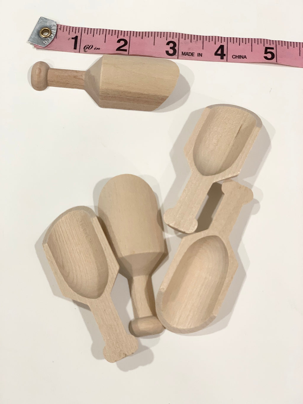 Five natural wooden sensory bin scoops and scoop with handle next to pink measuring tape on white surface